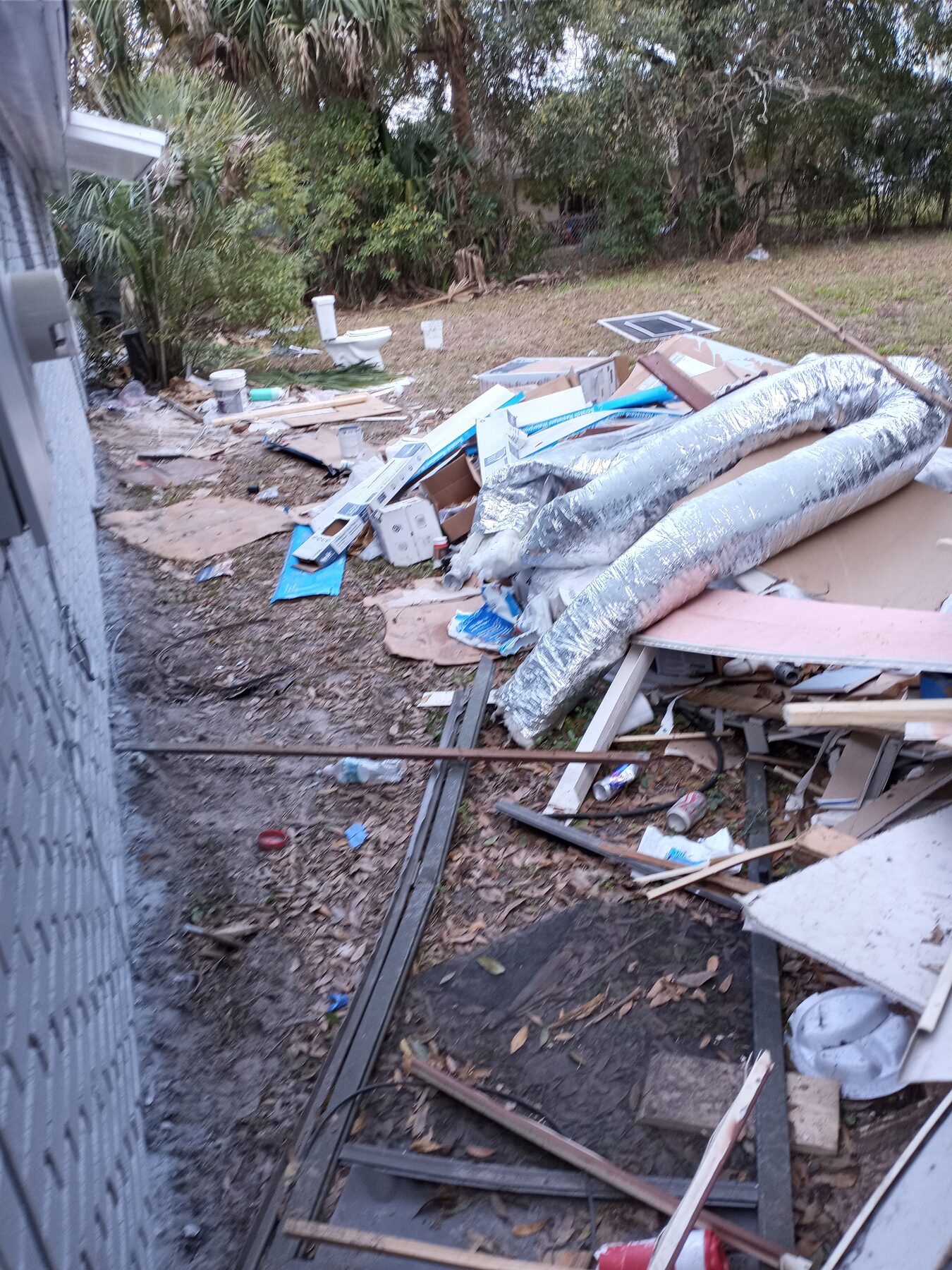 Back yard with toilet, ductwork, cardboard, and household debris scattered across grass before junk removal cleanout