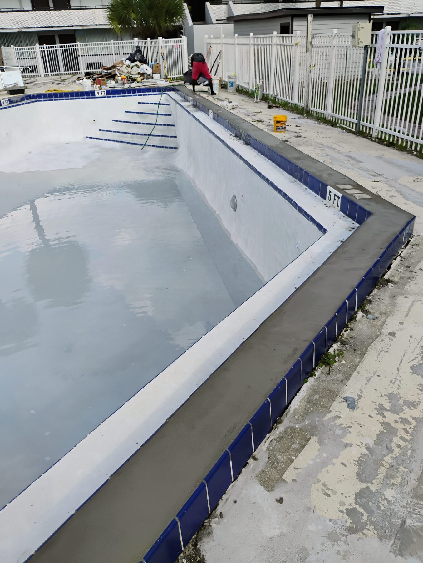 Commercial pool after replastering — fresh white plaster, new blue tile coping, bond beam finished, partially filled with water